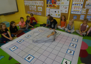Grupa dzieci siedzi wokół rozłożonej na dywanie macie do kodowania. Na macie ułożony jest zegar z tabliczek. Chłopiec leży na macie ułożony w literkę L wskazując tym samym godzinę 8.
